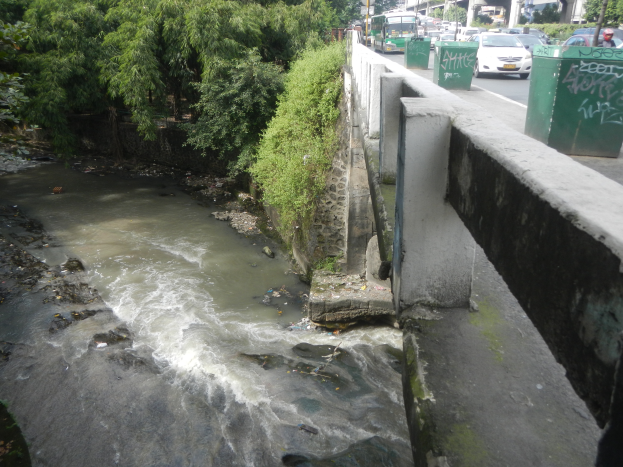Ein kleiner Bach fließt durch eine Stadtstraße neben einer Brücke, mit verschiedenen städtischen Elementen wie Gebäuden, Fahrzeugen, Menschen und Grünflächen im Vordergrund und dem Himmel im Hintergrund.