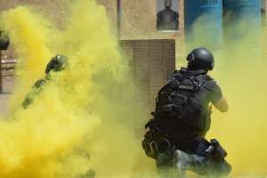 Zwei Polizisten in Schutzausrüstung mit gelbem Rauch aus dem Mund, mit Helmen und Schutzausrüstung, vor einer Wand mit einer Tafel und anderen Gegenständen, mit dem Boden unten sichtbar.