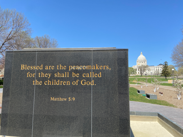 Ein Gedenkstein mit der Inschrift "Selig sind die Friedfertigen, denn sie sollen Kinder Gottes heißen" umgeben von Bäumen, Pflanzen, Gras, Laternenmasten und einem Gebäude unter einem klaren blauen Himmel.