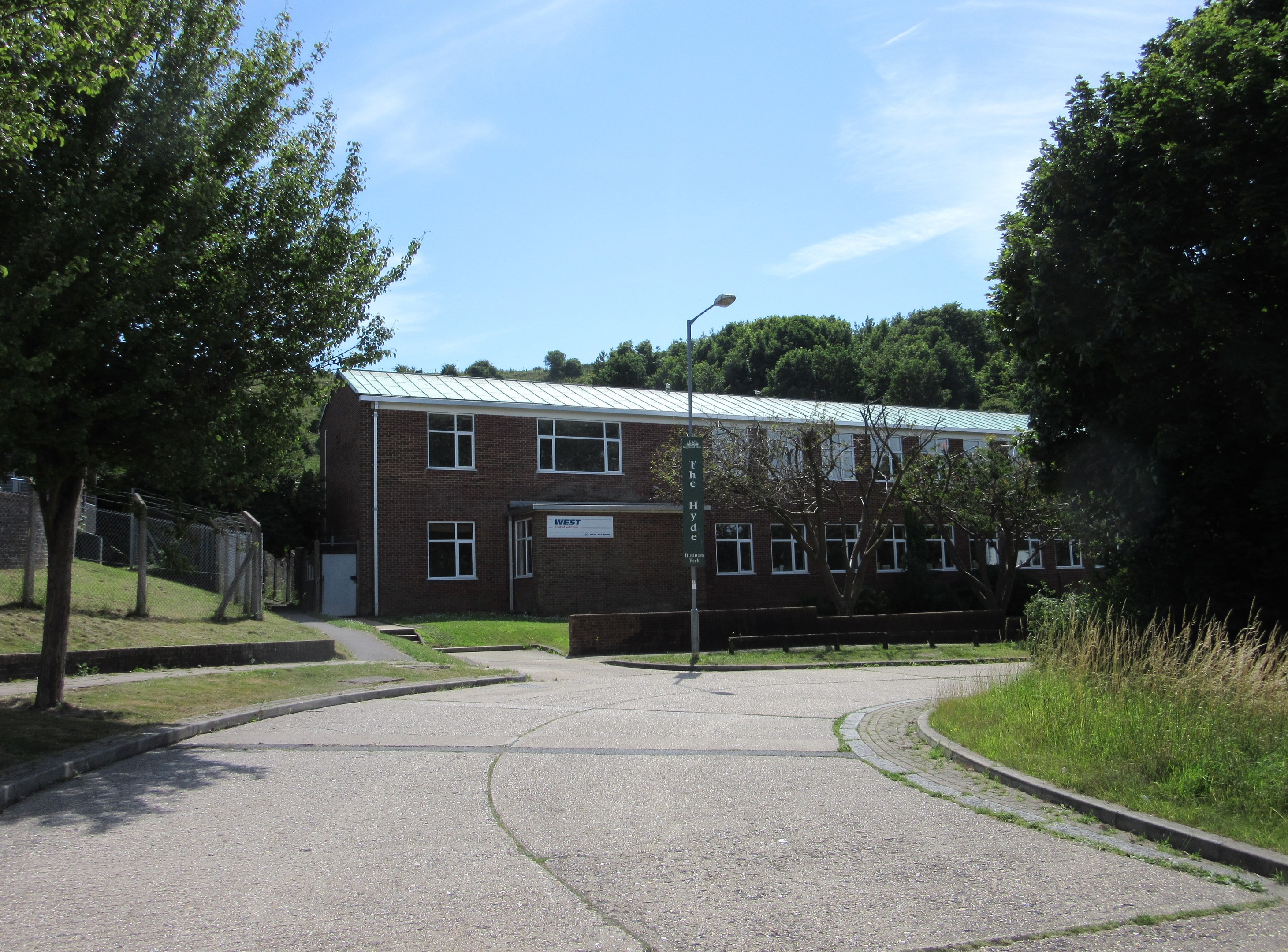Ein Schulgebäude mit Fenstern, ein Straßenschild, ein Schild, eine Baumgruppe, Gras, ein Zaun und ein bewölkter Himmel, umgeben von saftig grünem Gras in einem Park.