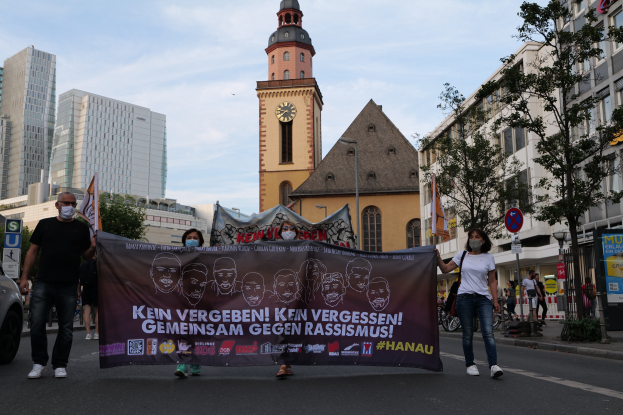 Eine Gruppe von Menschen mit Masken, die eine Fahne mit Text und Bildern halten, geht eine Straße entlang, mit einem geparkten Auto auf der linken Seite, Gebäuden, Bäumen, Schildern, Polen, einem Kirchturm und einem klaren blauen Himmel im Hintergrund.