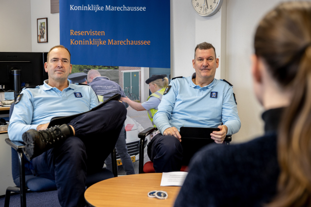 Zwei Polizeibeamte sitzen an einem Tisch mit Papieren und Gegenständen, sprechen mit einer Frau, mit einer Fahne, Uhr und Fotorahmen im Hintergrund und einem weiteren Tisch mit Monitor und Tastatur links daneben.