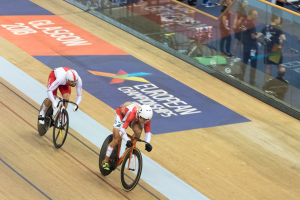 Zwei Männer mit Helmen fahren auf einer Bahn vor einem Publikum, mit einer Glaswand, auf der 'Europameisterschaften Glasgow 2018' steht und Menschen davor stehen und sitzen.