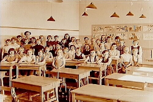 Ein Schwarz-Weiß-Foto eines Klassenzimmers mit Kindern auf Bänken und Stehenden, verstreuten Tischen und Stühlen, einer Tafel an der Wand und Deckenlampen, die den Raum erhellen.