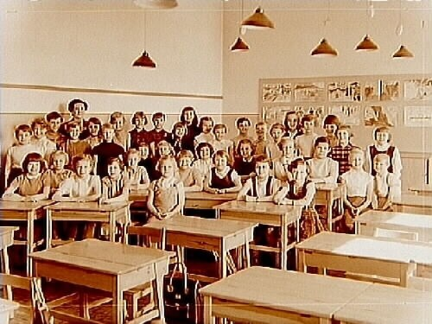 Ein Schwarz-Weiß-Foto eines Klassenzimmers mit Kindern auf Bänken und Stehenden, verstreuten Tischen und Stühlen, einer Tafel an der Wand und Deckenlampen, die den Raum erhellen.