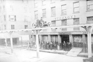 Altes Schwarz-Weiß-Foto eines Feuerwehrautos, das vor einem Gebäude mit einer Namenstafel geparkt ist, mit einer Person auf dem Fahrzeug und einer Gruppe von Menschen auf dem Gehweg sowie einem Hund in der Nähe.