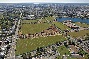 Luftaufnahme einer Stadt mit Gebäuden, Bäumen, Gras, Straßen mit Fahrzeugen, Wasser und Wolken, die einen großen zentralen Platz zeigt, der als Parkplatz vorgeschlagen ist.