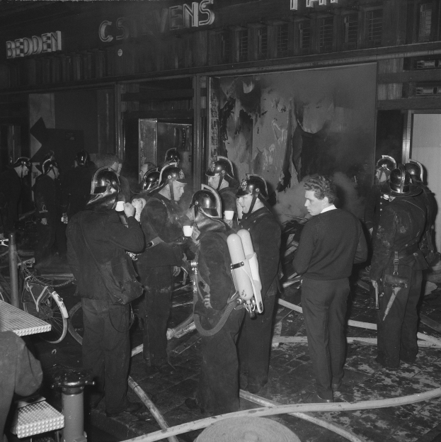 Schwarzes und weißes Foto von Feuerwehrleuten in Uniform mit Atemschutzgeräten neben einem Gebäude, einem Fahrrad und einem Tisch mit Gegenständen.
