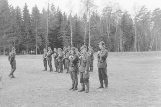 Schwarzes und weißes Bild einer Gruppe von Männern in militärischer Kleidung, die in einem Feld stehen, einige halten Gewehre, mit Bäumen und einem klaren Himmel im Hintergrund.