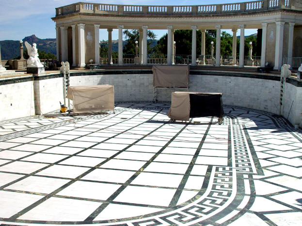 Ein Terrassenbereich mit drei Eisen-Tischen, einer Skulptur und Säulen, mit Laternen, Bäumen, einem Berg und dem Himmel im Hintergrund.