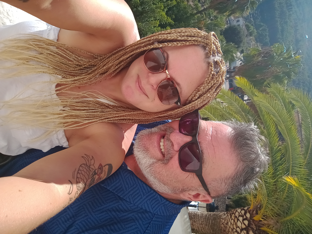 Ein Mann in einem blauen T-Shirt und eine Frau mit einer Sonnenbrille machen ein Selfie vor einer Palme, mit grünen Bäumen, Gebäuden und einem hellblauen Himmel im Hintergrund.