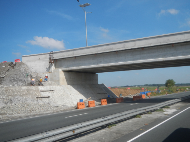 Autobahn mit einer Brücke darüber, ein darunter fahrendes Auto und Bauarbeiter am Straßenrand mit Absperrgittern, Steinen und einem Laternenmast; Bäume und bewölkter Himmel im Hintergrund.
