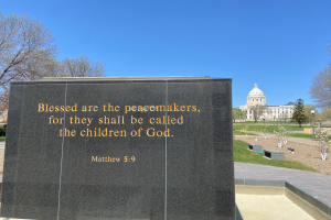 Ein Gedenkstein mit der Inschrift "Selig sind die Friedfertigen, denn sie sollen Kinder Gottes heißen" umgeben von Bäumen, Pflanzen, Gras, Laternenmasten und einem Gebäude unter einem klaren blauen Himmel.