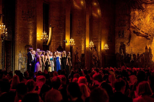 Eine Gruppe von Menschen auf einer beleuchteten Bühne im Royal Opera House in London mit bemalten Kulissen, die einer unten sitzenden Zuschauermenge gegenüberstehen.