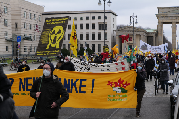 Eine große Gruppe von Menschen marschiert auf einer Straße, hält Schilder und Fahnen und demonstriert gegen Atomkraft in Deutschland, mit Fahrzeugen, Gebäuden, Laternenmasten, Bäumen und einem Tor im Hintergrund.