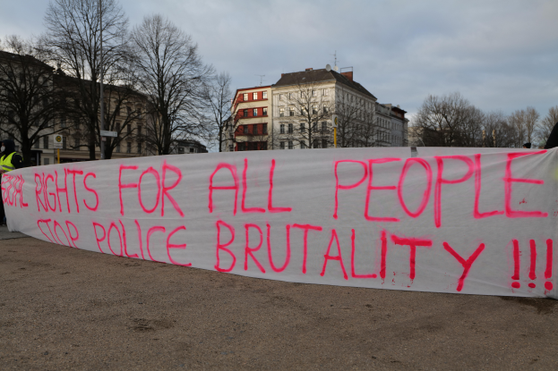Eine Gruppe von Menschen, die auf dem Boden stehen und ein Banner halten, auf dem 'Rechte für alle Menschen Stoppt Polizeigewalt' steht, mit einem Straßenschild, einem Schild, Bäumen, Gebäuden mit Fenstern und einem bewölkten Himmel im Hintergrund.