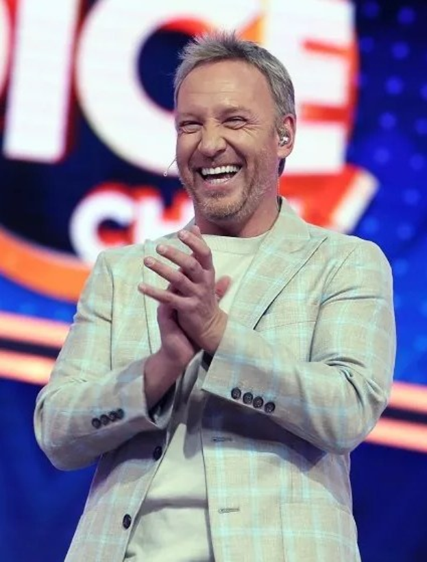 Ein Mann in einem Anzug, der auf einer Bühne klatscht, mit 'The Voice'-Plakat im Hintergrund.