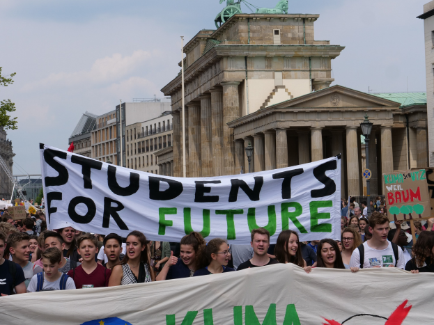 Gruppe von Studenten, die in Berlin marschieren und eine buntfarbene "Students for Future"-Fahne tragen, vor einer Kulisse aus Gebäuden, Bäumen und Himmel.