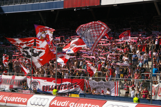 Eine große Menschenmenge in einem Stadion hält Fahnen und Banner, mit einem Metallzaun, einem Schild, einem Anzeigebildschirm und Deckenleuchten im Hintergrund.