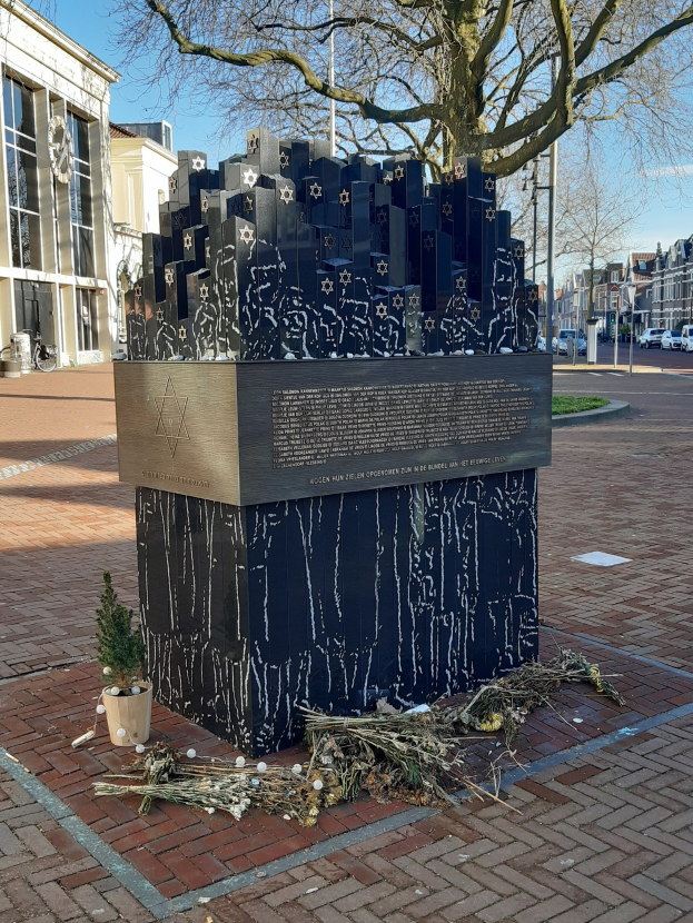 Denkmal in einem Stadtplatz für Holocaust-Opfer mit einem Baum davor, einem Topfgewächs, verstreuten trockenen Blättern und Gebäuden, Fahrzeugen und Bäumen im Hintergrund.