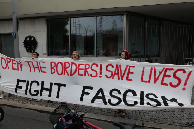 Eine Gruppe von Menschen hält ein Transparent mit der Aufschrift "Öffne die Grenzen, rette Leben, bekämpfe Faschismus" vor einem Gebäude mit Glasfenstern und einem Mast im Hintergrund, mit einem Fahrrad und einer Tasche auf der Straße.