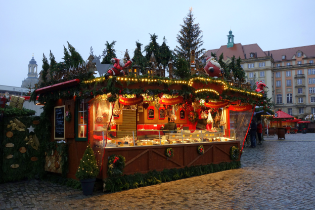 Ein Weihnachtsmarktstand auf einem Kopfsteinpflasterweg, umgeben von Gebäuden, geschmückten Bäumen und Menschen, mit sichtbarem Himmel im Hintergrund.
