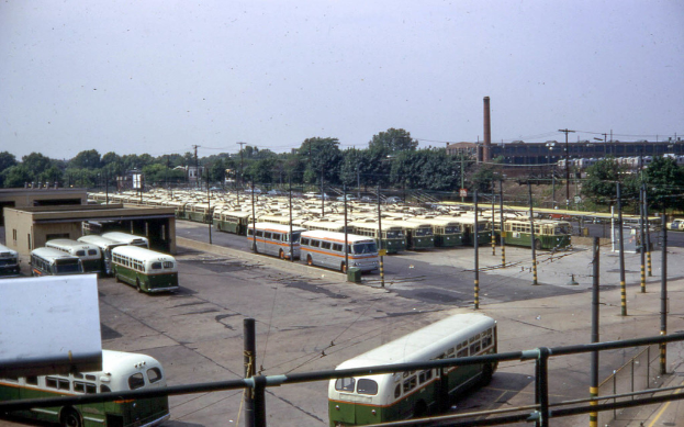 Mehrere Busse auf einer Straße mit Pfählen, Drähten, Gebäuden, Bäumen, einem Zaun und einem Rauchschlot, unter einem Himmel.