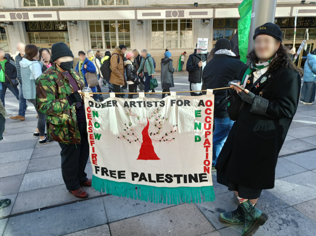 Eine Gruppe von Menschen, die auf dem Boden steht, einige halten ein Banner mit der Aufschrift 'Free Palestine', vor einem Gebäude mit Fenstern und einer Schautafel, mit einem Mast im Hintergrund.