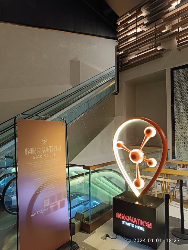 Großes Schild in einem Gebäude mit einer Rolltreppe im Hintergrund, flankiert von Tischen und Stühlen auf der rechten Seite und einer Tafel mit der Aufschrift "Innovation Starts Here" auf der linken Seite, die auf die HSTP20 Expo hinweist.