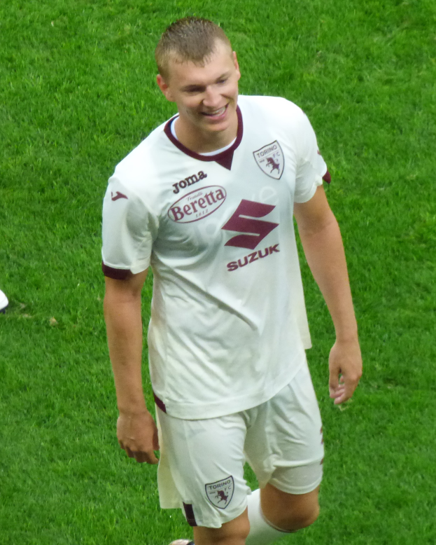 Ein Mann in weißem Hemd und Shorts lächelt beim Laufen auf einem Fußballfeld, trägt Schuhe.