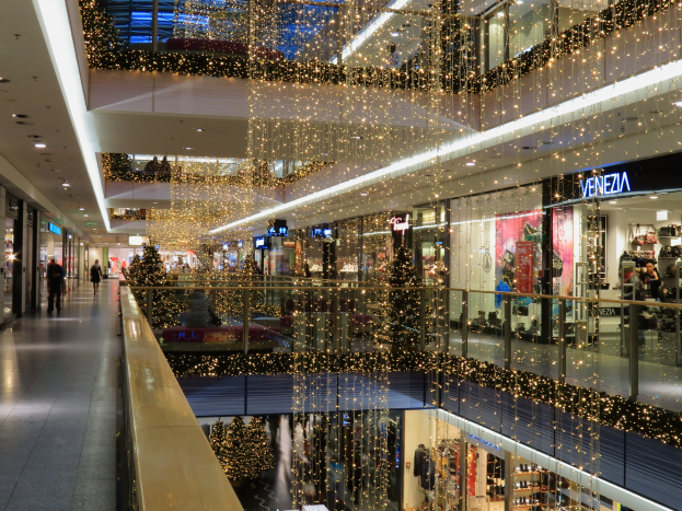 Ein festlich geschmücktes Einkaufszentrum mit Weihnachtsbeleuchtung, das Menschen zeigt, die durch die Geschäfte gehen, Schilder mit Namen, Glasgeländer und von der Decke hängende Lichter.
