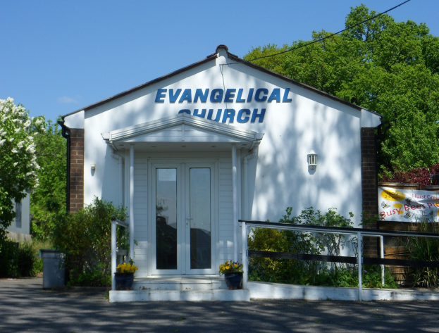 Evangelische Kirche mit Straße davor, umgeben von Bäumen und Pflanzen unter einem klaren blauen Himmel.