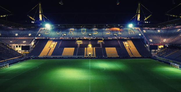 Ein Fuballfeld bei Nacht, erleuchtet von hellen Stadionlichtern, mit einigen Menschen auf den Trib├╝nen und saftig gr├╝nem Rasen.