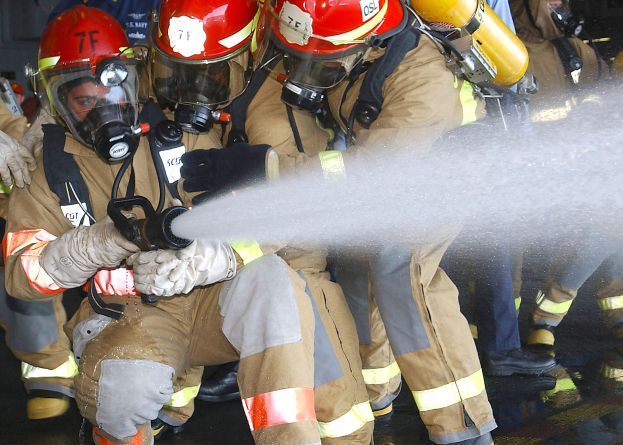 Feuerwehrleute in Schutzausrüstung sprühen Wasser aus einem Schlauch auf einen Boden in der Nähe einer Wand.