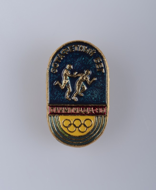 Ein runder Abzeichen mit dem Logo der Olympischen Spiele 1980, das zwei Läufer und den Text "Olympische Spiele" darüber zeigt.