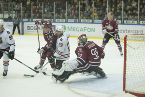 Gruppe von Menschen, die Hockey auf einem Eisstadion spielen, mit Torpfosten auf der rechten Seite, tragen Helme und halten Stöcke, Zuschauer auf Tribünen mit Bannern im Hintergrund.