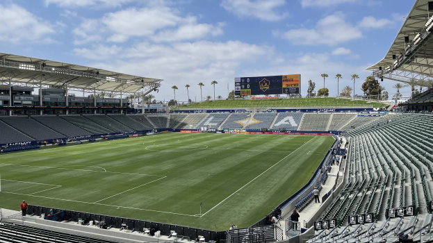 Ein Fußballfeld mit vielen Sitzplätzen, umgeben von Bäumen und einem bewölkten Himmel, das ein Spiel zwischen Los Angeles Galaxy und San Jose Earthquakes zeigt, mit Sitzenden und Stehenden Zuschauern und Anzeigetafeln mit den Namen der Mannschaften.