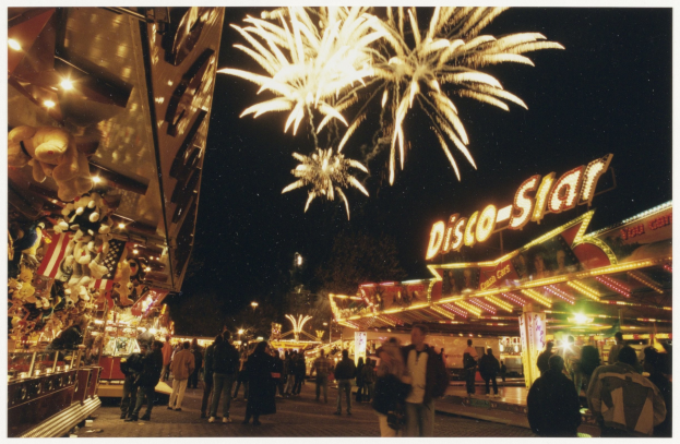 Eine Menschenmenge steht vor einer Karussellfahrt bei Nacht, beleuchtet von Lichtern und umgeben von Gebäuden mit Namensschildern, mit Bäumen und Feuerwerk im Hintergrund.