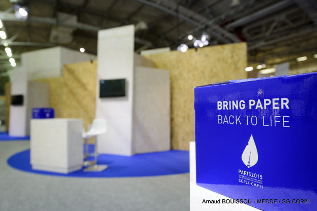 Eine Messestände mit einem blauen und weißen Schild "Bring Paper Back to Life" auf der rechten Seite, mit unscharfen Stühlen, Tischen, einem Fernseher und Deckenlampen im Hintergrund.