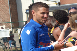 Ein Mann in einem blauen Hemd signiert Autogramme für eine Gruppe von Menschen, einige tragen Mützen und Schutzbrillen, mit einem Gebäude, Maschendrahtzaun und einem Fahrrad im Hintergrund.