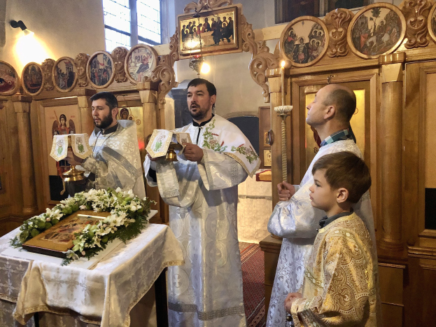 Eine Gruppe von Menschen steht um einen Tisch in einer Kirche, hält Bücher in den Händen, mit einem Tuch und einem Blumenstrauß auf dem Tisch, Rahmen an der Wand, Lampen, einem Fenster und einem hölzernen Objekt im Hintergrund.