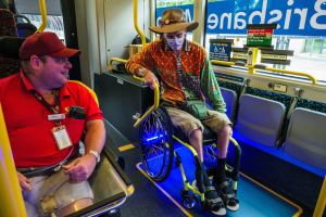 Zwei Männer im Rollstuhl auf einem Bus, einer trägt ein rotes T-Shirt und eine rote Mötte, der andere ein orangefarbenes Hemd und einen braunen Hut, beide tragen Masken und Ausweise, mit gelben Stangen und Glasfenstern, durch die man Bäume, Gebäude und den Himmel sieht.