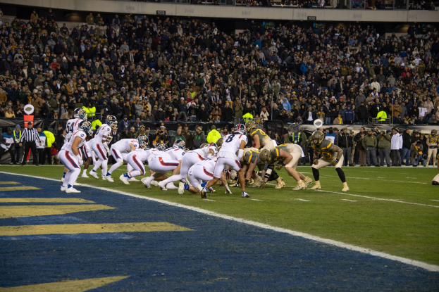 Ein Fußballspiel im Gange mit Spielern in Sportbekleidung und Helmen auf dem Boden, von einem Stadionplatz aus betrachtet.