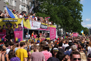 Eine große Menge marschiert auf einer Straße während des Christopher Street Day 2019 in Berlin, viele tragen Mützen und Schutzbrillen, einige halten Fahnen, mit Bannern, Laternenmasten, Bäumen und Gebäuden im Hintergrund bei bewölktem Himmel.