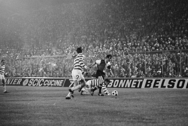 Eine Gruppe von Männern, die Fußball auf einem Feld spielen, mit Zuschauern auf den Tribünen und einem Banner mit der Aufschrift "Celtic v West Bromwich Albion - FA Cup Third Round", in Schwarz-Weiß.