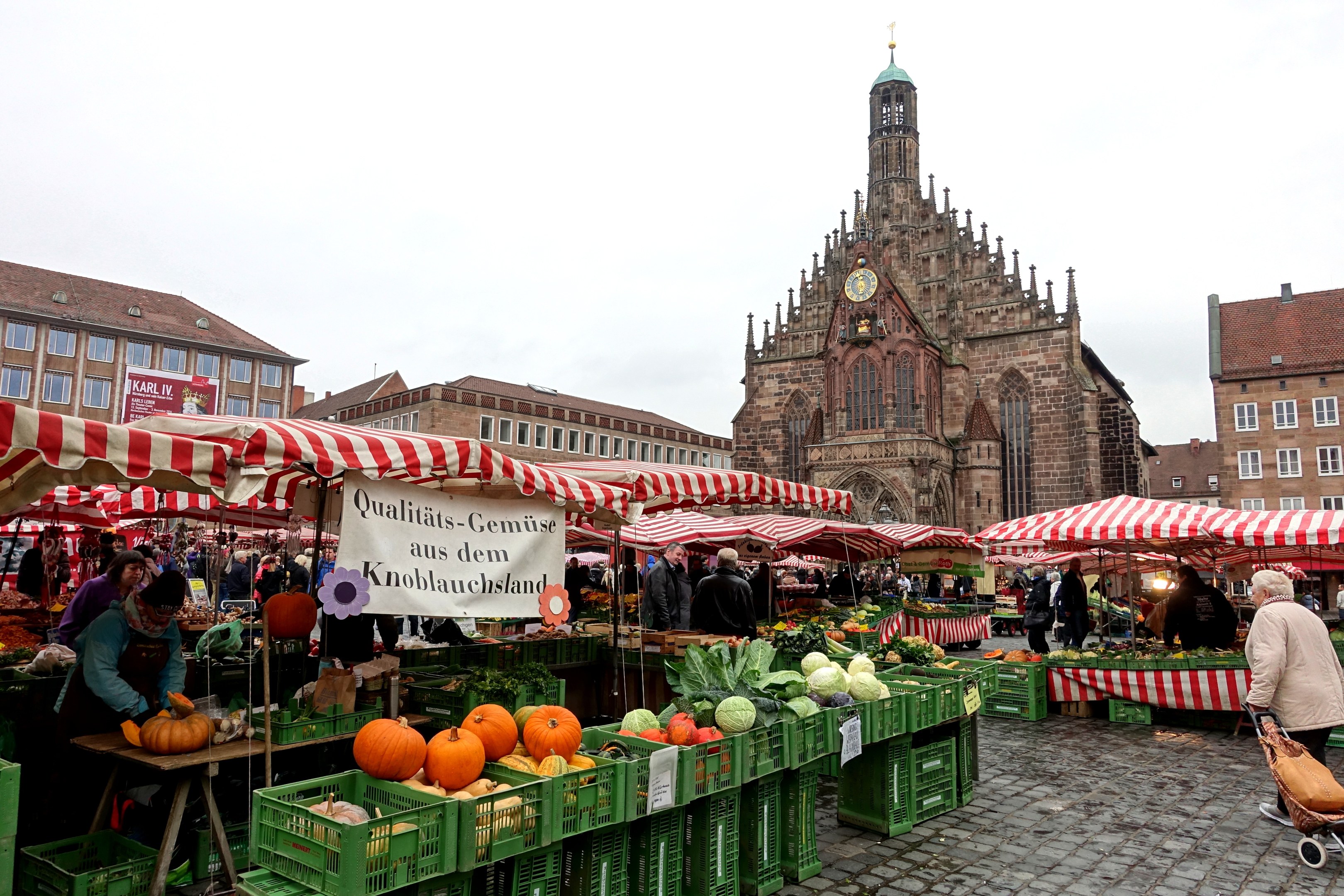 Ein belebter Markt in Nürnberg, Deutschland, mit farbenfrohen Obst und Gemüse, Menschen mit Taschen und um den Markt aufgestellte Zelte, sowie Gebäude und ein Uhrenturm im Hintergrund unter einem sichtbaren Himmel.