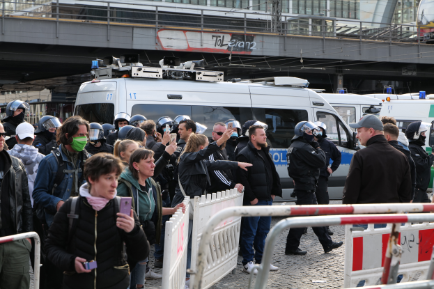 Eine Gruppe von Menschen steht vor einer Reihe von Polizeiwagen in Berlin, einige tragen Helme und halten Telefone, mit Barrieren im Vordergrund und einer Brücke mit Geländern und Gebäuden im Hintergrund.