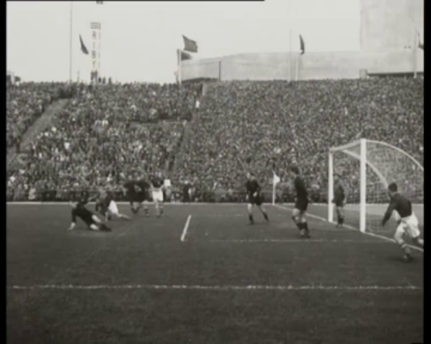 Ein Schwarz-Weiß-Foto von einem WM-Finale 1958 zwischen Manchester United und Liverpool, mit Spielern auf dem Feld, einem Torpfosten auf der rechten Seite, Zuschauern auf den Rängen und Fahnen im Hintergrund.