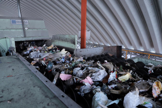 Großer Müllberg auf einem Förderband in einer Recyclinganlage mit verstreutem Müll und einem Metalldach darüber.