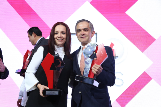 Zwei Menschen, die auf einer Bühne lächeln und Trophäen halten, mit einem sichtbaren Bildschirm im Hintergrund.